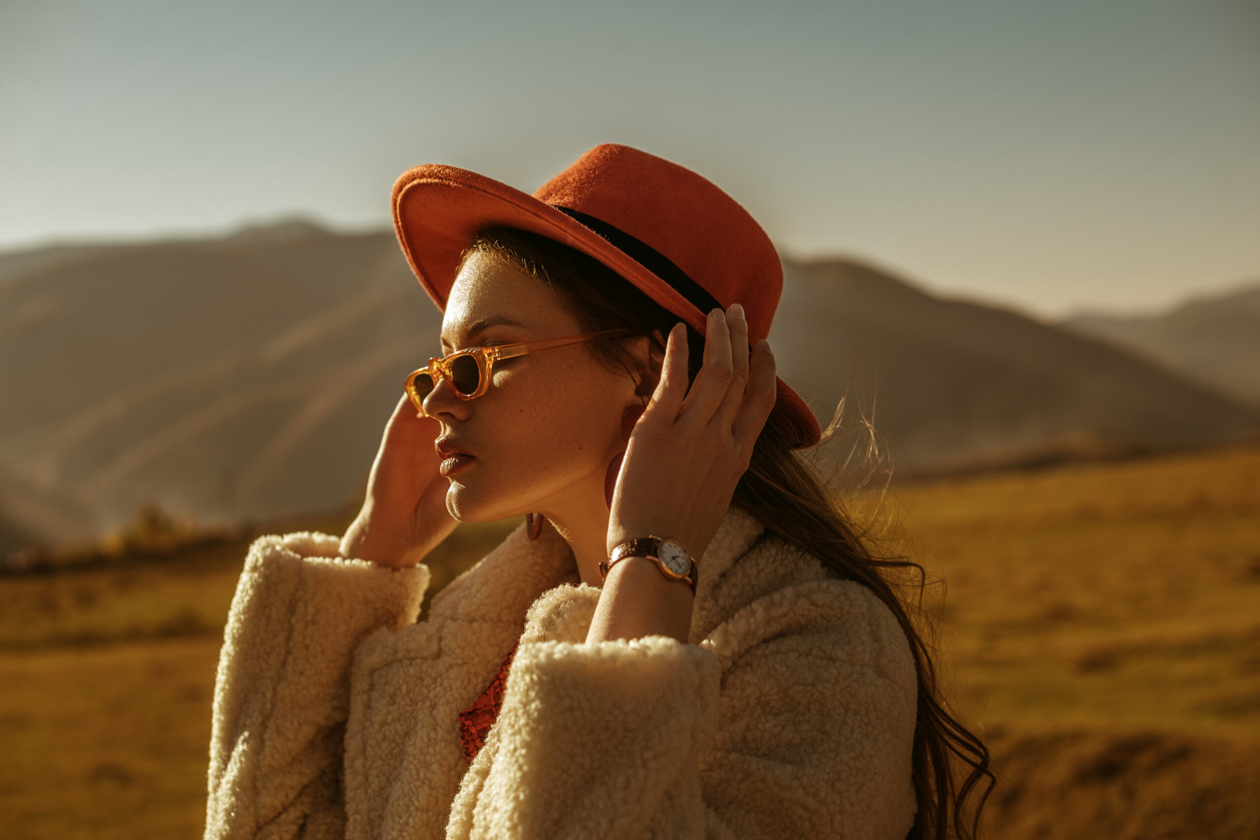 Outdoor close up fashion portrait of young beautiful confident brunette woman wearing stylish orange hat, sunglasses, wrist watch, faux fur coat, posing in mountain landscape. Copy, empty space